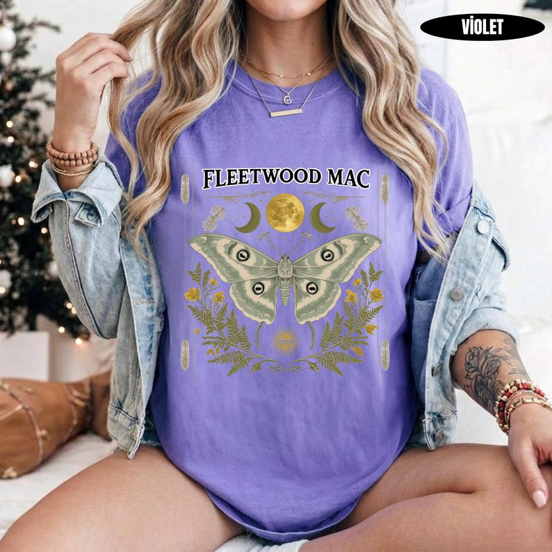 Może przedstawiać: Fioletowy t-shirt z kr&oacute;tkim rękawem z napisem Fleetwood Mac nad grafiką przedstawiającą dużą ćmę, ż&oacute;łty księżyc i zielone liście paproci. Koszulka jest założona pod jasnoniebieską kurtkę dżinsową z metalowymi guzikami i widocznymi szwami.