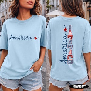 May include: A light blue short-sleeve t-shirt featuring the word America in dark blue script with a red star on the front. The back displays the word America and the number 1775 in a vertical design with red, white, and blue flag patterns.