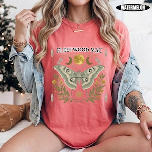 Może przedstawiać: Koralowy t-shirt z grafiką przedstawiającą ćmę, pełnię księżyca, p&oacute;łksiężyce i liście paproci. Nad ćmą znajduje się napis FLEETWOOD MAC. Koszulka jest założona pod jasnoniebieską kurtkę dżinsową z długimi rękawami i metalowymi guzikami.