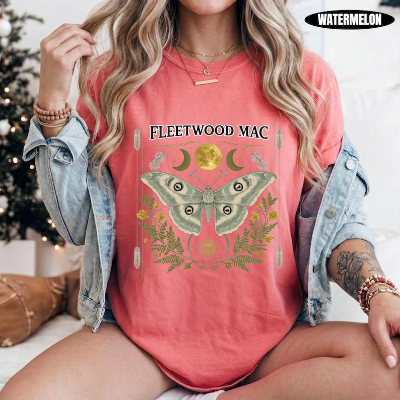 Może przedstawiać: Koralowy t-shirt z grafiką przedstawiającą ćmę, pełnię księżyca, p&oacute;łksiężyce i liście paproci. Nad ćmą znajduje się napis FLEETWOOD MAC. Koszulka jest założona pod jasnoniebieską kurtkę dżinsową z długimi rękawami i metalowymi guzikami.