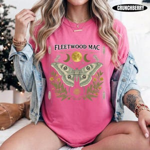 Może przedstawiać: Jaskrawor&oacute;żowy t-shirt z kr&oacute;tkim rękawem z nadrukiem graficznym przedstawiającym dużą ćmę, ż&oacute;łty księżyc i zielone liście paproci. Napis FLEETWOOD MAC jest nadrukowany na czarno nad ćmą.