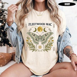 May include: An ivory t-shirt featuring a graphic print with the text FLEETWOOD MAC, a large moth, a golden moon, crescent moons, and green fern leaves. The design includes a sunburst symbol at the bottom and vertical line accents on the sides.
