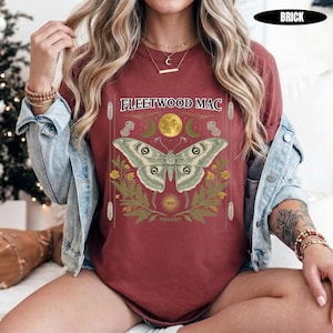 Może przedstawiać: Ceglastoczerwony t-shirt z kr&oacute;tkim rękawem z napisem FLEETWOOD MAC nad grafiką przedstawiającą dużą ćmę, ż&oacute;łty księżyc i zielone liście. Koszulka założona pod jasnoniebieską kurtkę dżinsową, z bransoletkami z koralik&oacute;w na obu nadgarstkach.