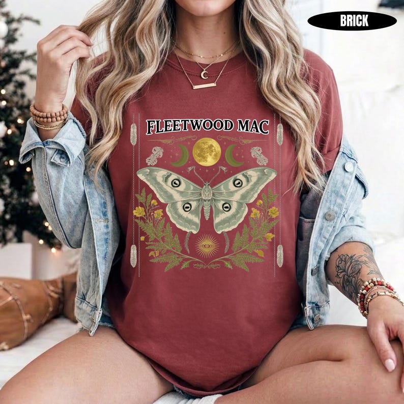 Może przedstawiać: Ceglastoczerwony t-shirt z kr&oacute;tkim rękawem z napisem FLEETWOOD MAC nad grafiką przedstawiającą dużą ćmę, ż&oacute;łty księżyc i zielone liście. Koszulka założona pod jasnoniebieską kurtkę dżinsową, z bransoletkami z koralik&oacute;w na obu nadgarstkach.