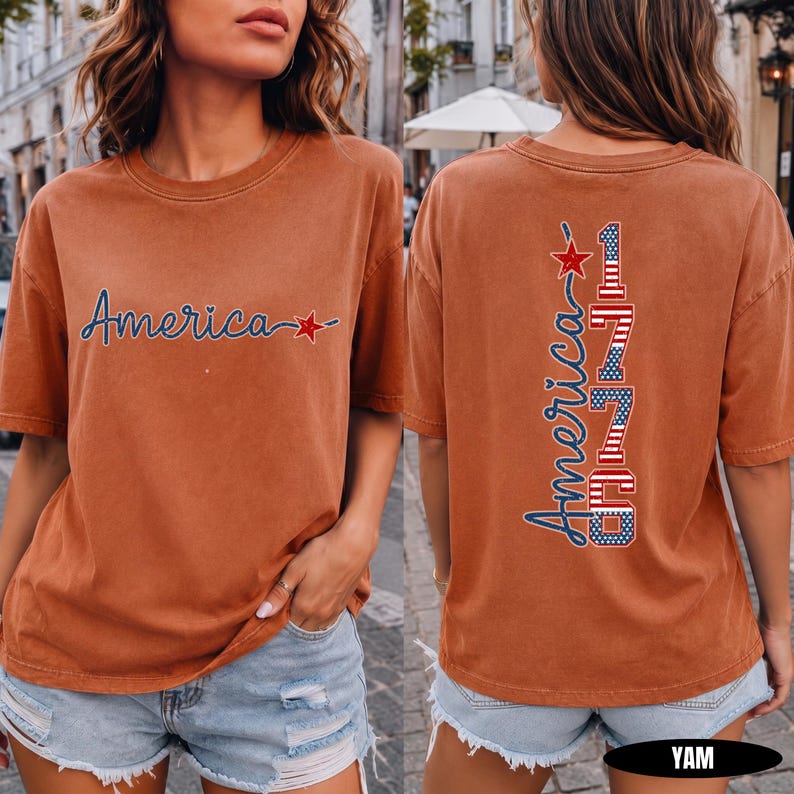May include: A rust-colored short-sleeve t-shirt featuring the word America in blue script with a red star on the front. The back displays the word America vertically in blue script next to the number 1776 in red, white, and blue flag patterns.