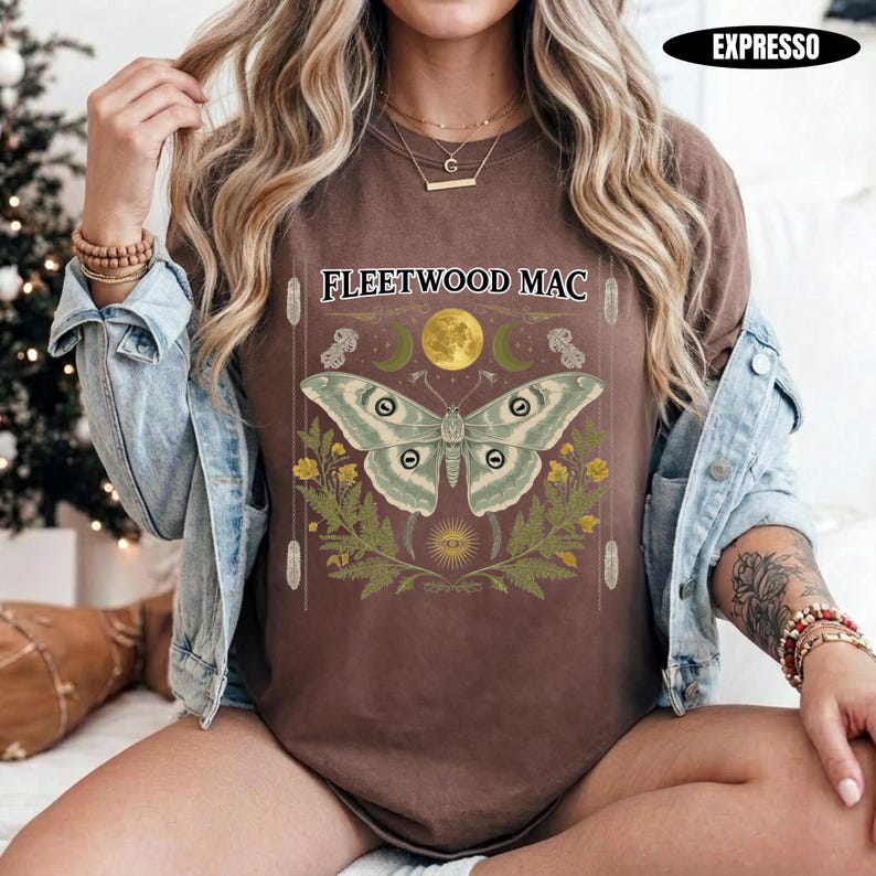 Może przedstawiać: Brązowy t-shirt z napisem Fleetwood Mac nad ilustracją dużej ćmy, ż&oacute;łtym księżycem i zielonymi liśćmi. Wz&oacute;r zawiera małe symbole niebieskie i pionowe linie. T-shirt jest noszony pod jasnoniebieską kurtką dżinsową.