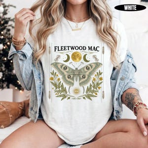 Może przedstawiać: Biały t-shirt z nadrukiem graficznym zawierającym napis FLEETWOOD MAC, dużą ćmę, pełnię księżyca, p&oacute;łksiężyce, liście paproci, ż&oacute;łte kwiaty oraz symbol oka. Koszulka jest założona pod jasnoniebieską kurtkę dżinsową z długimi rękawami.