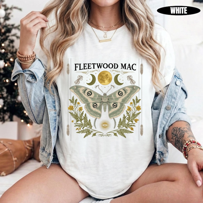 Może przedstawiać: Biały t-shirt z nadrukiem graficznym zawierającym napis FLEETWOOD MAC, dużą ćmę, pełnię księżyca, p&oacute;łksiężyce, liście paproci, ż&oacute;łte kwiaty oraz symbol oka. Koszulka jest założona pod jasnoniebieską kurtkę dżinsową z długimi rękawami.