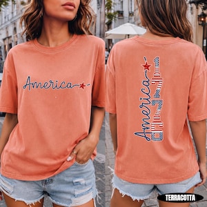 May include: A terracotta-colored short-sleeve t-shirt featuring the word America in blue cursive script with a red star on the front. The back displays the word America vertically next to the number 1776, both filled with a red, white, and blue flag pattern.
