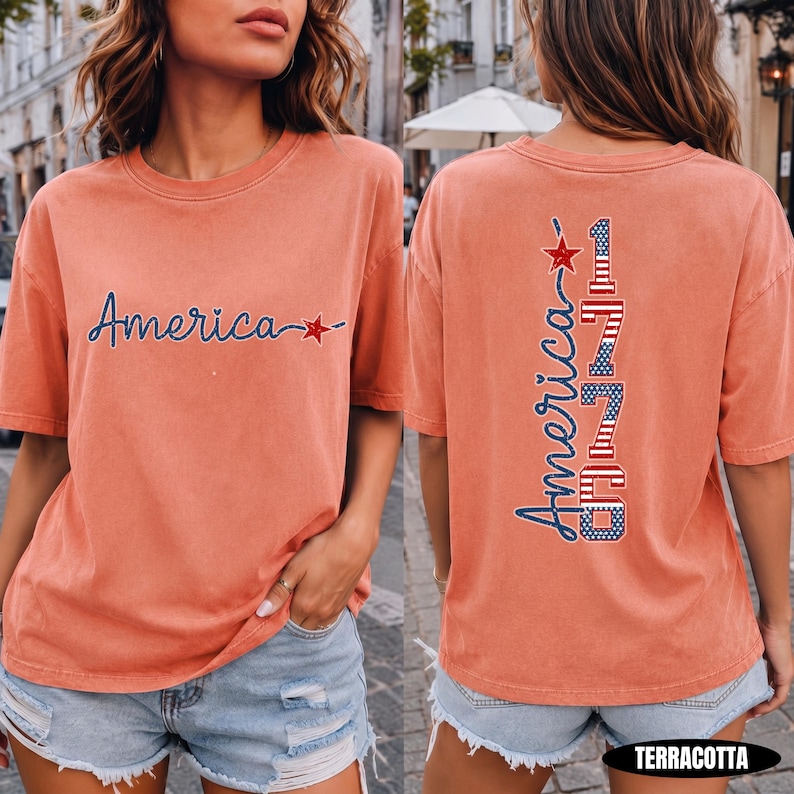 May include: A terracotta-colored short-sleeve t-shirt featuring the word America in blue cursive script with a red star on the front. The back displays the word America vertically next to the number 1776, both filled with a red, white, and blue flag pattern.