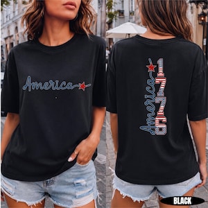 May include: A black short-sleeve t-shirt featuring the word America in blue script with a red star on the front. The back displays the word America vertically next to the number 1776, styled with red, white, and blue American flag patterns.