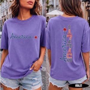 May include: A violet short-sleeved t-shirt featuring the word America in dark blue cursive script with a red star on the front. The back displays the word America vertically alongside the number 1776, both filled with a red, white, and blue flag pattern.