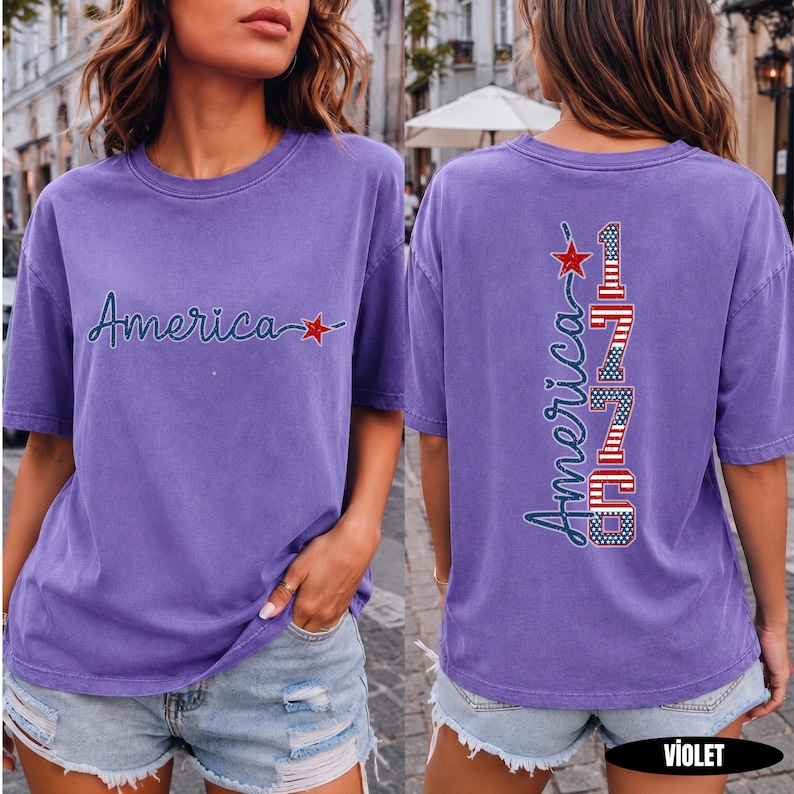 May include: A violet short-sleeved t-shirt featuring the word America in dark blue cursive script with a red star on the front. The back displays the word America vertically alongside the number 1776, both filled with a red, white, and blue flag pattern.