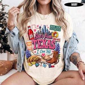 May include: An ivory t-shirt featuring a graphic print with the text "She's From TEXAS I Can Tell," a Dallas and San Antonio road sign, a Big Red soda bottle, a Whataburger cup, a cowboy boot, a cowboy hat, tacos, and a Texas state outline.