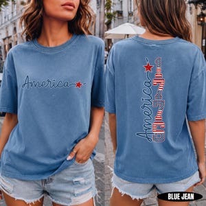 May include: A blue short-sleeve t-shirt featuring the word America in cursive script with a red star on the front. The back displays the word America vertically alongside the number 1776, both filled with a red, white, and blue flag pattern.