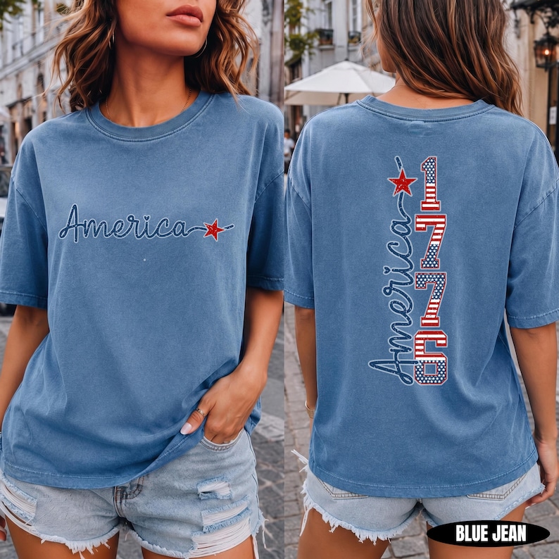 May include: A blue short-sleeve t-shirt featuring the word America in cursive script with a red star on the front. The back displays the word America vertically alongside the number 1776, both filled with a red, white, and blue flag pattern.