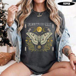 Może przedstawiać: Ciemnoszary t-shirt z nadrukiem graficznym przedstawiającym dużą ćmę nad ż&oacute;łtymi kwiatami i paprociami. Nad ćmą znajduje się napis FLEETWOOD MAC, ż&oacute;łty księżyc oraz p&oacute;łksiężyce. Koszulka jest założona pod jasnoniebieską kurtkę dżinsową.
