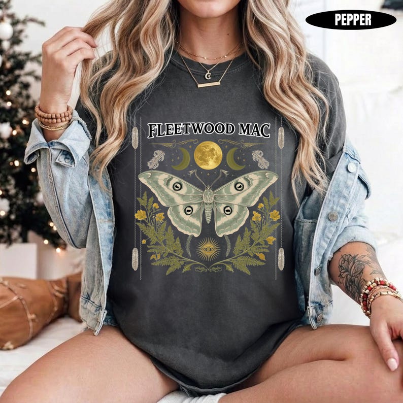 Może przedstawiać: Ciemnoszary t-shirt z nadrukiem graficznym przedstawiającym dużą ćmę nad ż&oacute;łtymi kwiatami i paprociami. Nad ćmą znajduje się napis FLEETWOOD MAC, ż&oacute;łty księżyc oraz p&oacute;łksiężyce. Koszulka jest założona pod jasnoniebieską kurtkę dżinsową.
