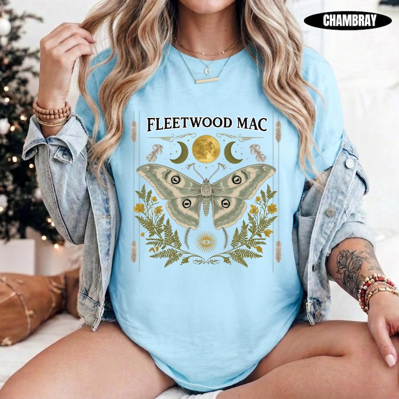 Może przedstawiać: Jasnoniebieski t-shirt z nadrukiem graficznym przedstawiającym dużą ćmę umieszczoną nad napisem FLEETWOOD MAC. Wz&oacute;r zawiera złoty księżyc, liście paproci oraz niebiańskie symbole w odcieniach zieleni, złota i brązu na przodzie koszulki.