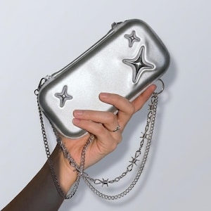 Op de afbeelding: Zilveren rechthoekige crossbodytas met sterversieringen. De tas heeft een kettingriem met een prikkeldraadontwerp. De tas wordt door een hand tegen een witte achtergrond gehouden.