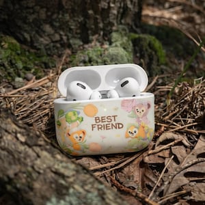 May include: White wireless earbuds in an open case with a colorful design. The case features illustrations of cartoon characters and the words "BEST FRIEND". The earbuds are resting on a bed of leaves and twigs.