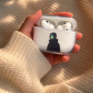 May include: Clear protective case for wireless earbuds, featuring a graphic of a character in a black cloak with green accents. The case is open, revealing white earbuds inside. The case is held in a person's hand.
