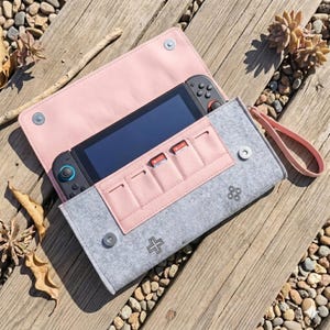 Puede incluir: Una funda de fieltro gris y rosa para una consola de juegos, abierta para revelar la consola y los cartuchos de juego. La funda tiene un interior rosa y una correa de muñeca. La parte delantera de la funda presenta un símbolo más e iconos de botones de control.