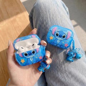 Può includere: Due custodie blu per AirPods con un design a personaggio dei cartoni animati. Una custodia è aperta, rivelando auricolari bianchi. Un piccolo portachiavi con la figura di Stitch è attaccato a una custodia. Le custodie sono decorate con stelle.