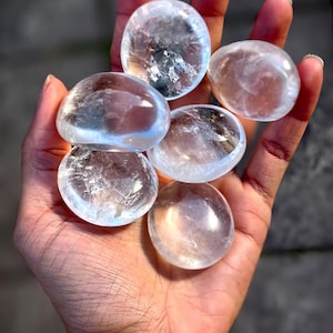 Pode incluir: Uma mão segura seis pedras de palma de quartzo transparente. As pedras são lisas, arredondadas e translúcidas, com padrões internos sutis. As pedras variam ligeiramente em tamanho e forma, em um fundo desfocado.