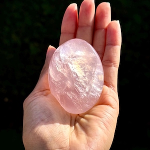 Puede incluir: Una piedra de cuarzo rosa lisa y ovalada, sostenida en una mano. La piedra es de un color rosa suave con áreas translúcidas, que capturan la luz. El fondo es un verde oscuro borroso, enfatizando el color y la textura de la piedra.