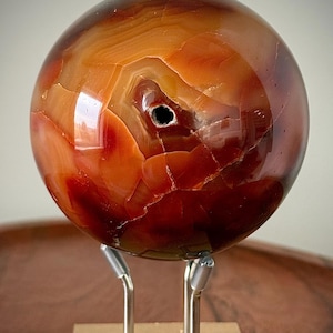 Peut inclure: Une sphère en cornaline polie présentant des motifs en bandes orange foncé, rouges et brunes avec une petite ouverture naturelle sombre près du centre. La pierre est exposée sur un support en métal argenté à trois branches et base carrée.