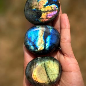 May include: Three polished labradorite stones stacked vertically. The top stone displays iridescent blue, yellow, and pink flashes. The middle stone features a vibrant blue and gold sheen. The bottom stone shows a soft green and yellow glow.