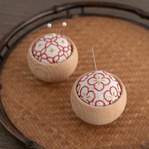 Puede incluir: Dos cojines de alfiler de madera con una parte superior de tela estampada floral y una base de madera. La tela es blanca con un diseño floral rojo. Las agujas están insertadas en los cojines. Los cojines de alfiler están en una bandeja tejida.