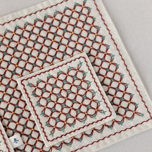 Può includere: Un set di sottobicchieri e tovaglietta in lino ricamato. Il tessuto bianco sporco è decorato con un motivo geometrico in filo rosso, verde acqua e oro. I bordi sono rifiniti con una cucitura rossa decorativa.