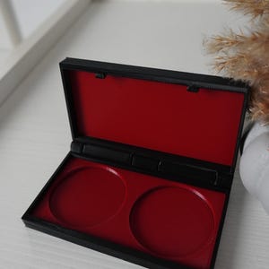 Puede incluir: Un estuche de maquillaje negro rectangular con interior rojo. El estuche está abierto, revelando dos huecos circulares. El fondo es una superficie blanca con un elemento decorativo.