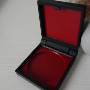 Pode incluir: Uma caixa compacta quadrada preta com interior vermelho. A caixa está aberta, revelando uma reentrância circular. A caixa está sobre uma superfície branca.