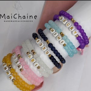 May include: A selection of beaded bracelets in shades of yellow, pink, white, navy, light blue, and purple. Each bracelet has gold details and white letter beads spelling out names or phrases. The brand name "MaiChaine" is visible.