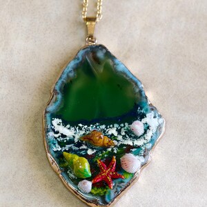Peut inclure: Collier avec une chaîne dorée et un pendentif en agate verte. Le pendentif représente une scène océanique miniature avec des coquillages, une étoile de mer et un bord doré.