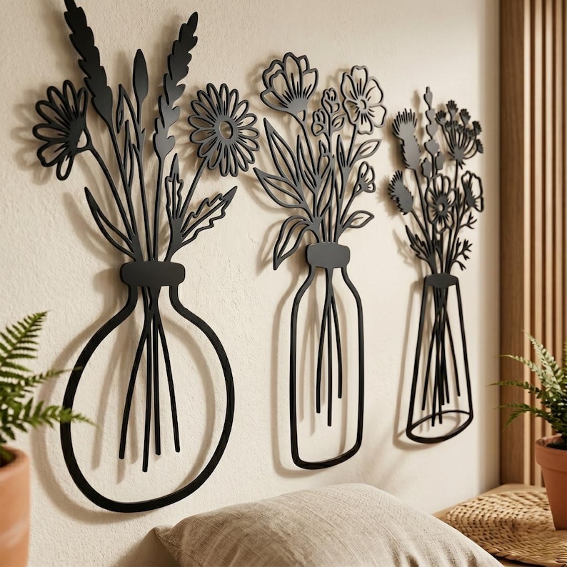May include: Three black metal wall art pieces depicting stylized flowers in vases. The vases vary in shape, with one round, one rectangular, and one triangular. The floral arrangements include daisies, leaves, and other botanical elements. The art is mounted on a neutral-colored wall.