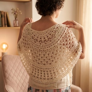 May include: A cream-colored, semi-circular crochet shawl featuring an intricate, openwork lace pattern with a scalloped edge. The shawl is draped over the shoulders, showcasing a detailed, repeating geometric design that radiates from the center.