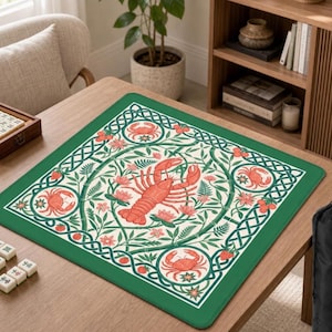 May include: A square game mat featuring a central orange lobster surrounded by pink flowers, green ferns, and berries. The design includes four circular frames with orange crabs at the corners, all enclosed by a dark green Celtic knot border.