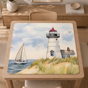 May include: A square table mat featuring a watercolor illustration of a white lighthouse with a red roof, a sailboat on blue water, and tall green grass in the foreground. The scene includes a light blue sky with soft clouds and flying birds.