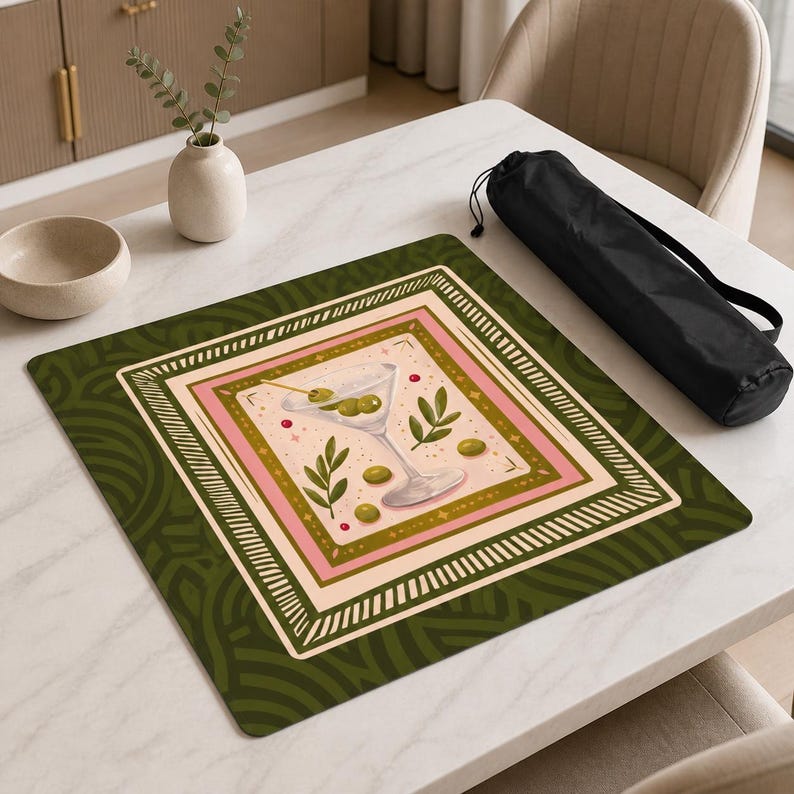 May include: A square mat featuring a central illustration of a martini glass with olives, surrounded by a pink and cream border and a dark green patterned background. The design includes small green leaves and red dots, presented with a black carrying case.