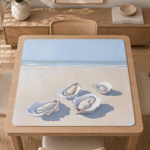 May include: A square table mat featuring a printed illustration of four open oyster shells resting on a sandy beach. The background depicts a calm shoreline with a light blue sky meeting the horizon, creating a serene coastal scene on the mat.