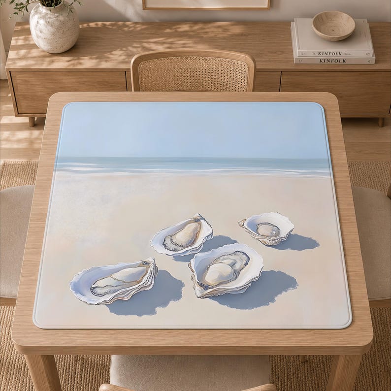 May include: A square table mat featuring a printed illustration of four open oyster shells resting on a sandy beach. The background depicts a calm shoreline with a light blue sky meeting the horizon, creating a serene coastal scene on the mat.