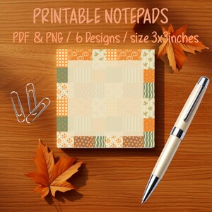 May include: A square notepad featuring a patchwork border of orange, green, and cream patterns with pumpkins, stripes, and dots. The notepad is accompanied by three silver metal paper clips and a cream-coloured ballpoint pen with silver accents.