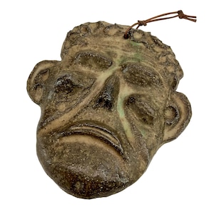 Könnte beinhalten: Ein handgefertigter Keramik-Wandschmuck in Form eines menschlichen Gesichts mit einer texturierten, gesprenkelten braunen und beigen Glasur. Das Gesicht zeigt tief liegende Augen, eine markante Nase und einen herabgezogenen Mund.