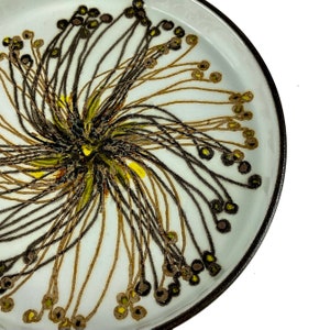 Può includere: Un piatto in ceramica bianca con bordo nero e un motivo floreale centrale. Il design presenta pennellate marroni, nere, gialle e verdi che si irradiano dal centro. Il piatto è rotondo.