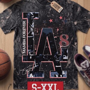 May include: Black and gray graphic t-shirt featuring the text "MAMBA FOREVER" and the letters "LA" with images of a basketball player. The shirt includes the text "S-XXL" and "MAMBA DAY EDITION".