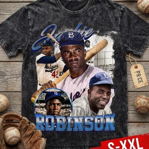 May include: A charcoal gray t-shirt featuring a collage of Jackie Robinson images, including portraits and action shots. The design includes the name "Jackie" in blue script and the word "Robinson" in blue block letters. The shirt is available in sizes S-XXL.